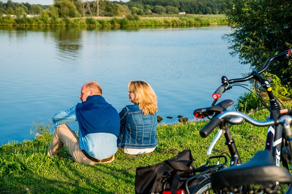 Frau und Mann sitzen am Ufer der Maas und ruhen sich während ihrer Fahrradtour durch die Gemeinde Leudal aus