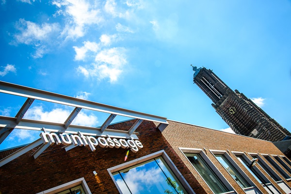 Ingang van winkelcentrum Muntpassage in Weert, met de toren van de Sint-Martinuskerk op de achtergrond tegen een blauwe lucht met wolken