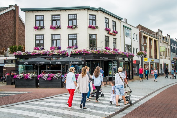 Shoppers steken over in het centrum van Weert, langs een terras met bloemen en diverse winkels op de achtergrond.